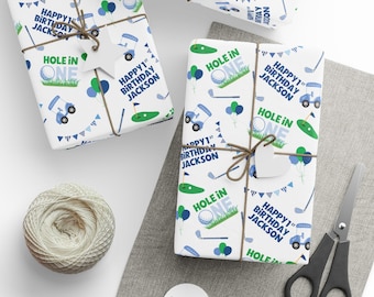 Op de afbeelding: Wit inpakpapier met een herhalend golfpatroon met blauwe golfkarretjes, groene putting greens, golfballen en ballonnen. De tekst luidt "Hole in One" en "Happy 1st Birthday Jackson" in blauwe en groene inkt.