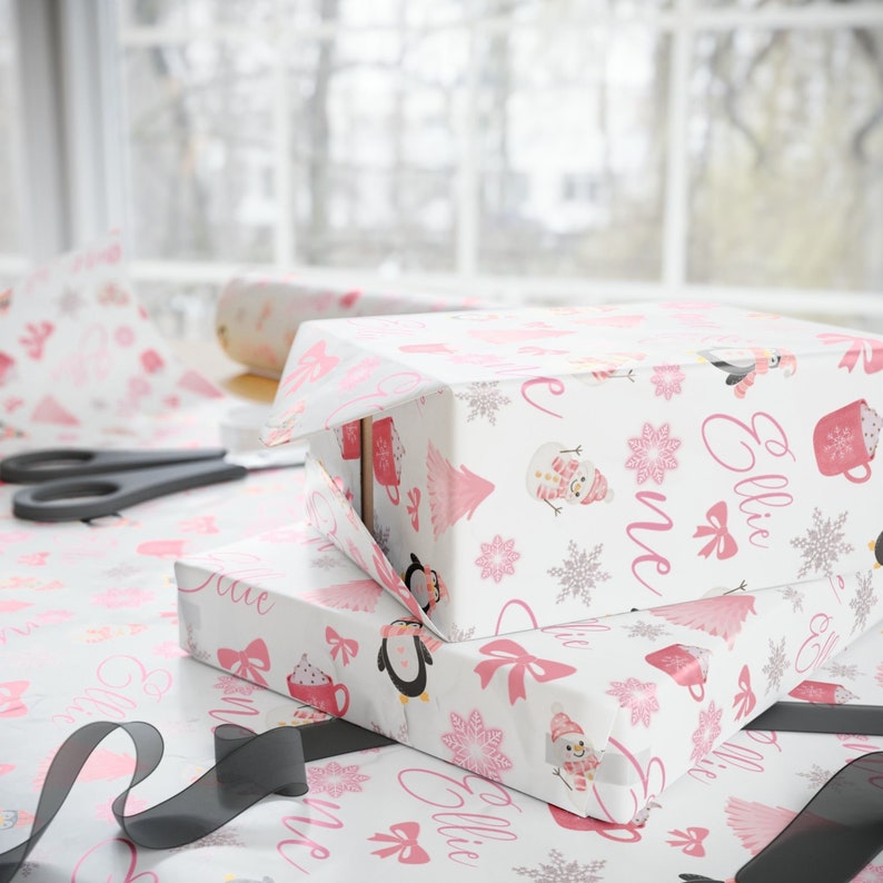 May include: Two white gift boxes with pink and white patterned wrapping paper. The wrapping paper features a snowman, penguins, snowflakes, and the name "Ellie" repeated on the paper.
