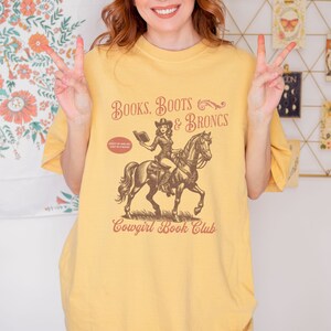 May include: Yellow t-shirt with a vintage illustration of a cowgirl riding a horse and reading a book. The text on the shirt reads "Books, Boots & Broncs" and "Cowgirl Book Club".