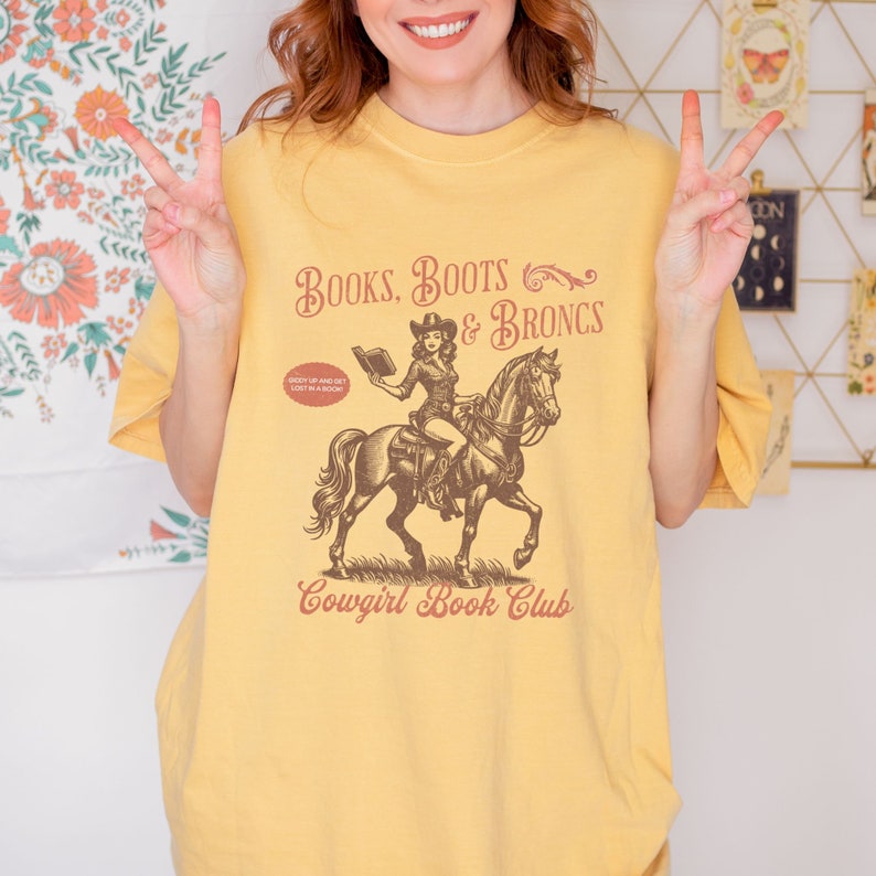 May include: Yellow t-shirt with a vintage illustration of a cowgirl riding a horse and reading a book. The text on the shirt reads "Books, Boots & Broncs" and "Cowgirl Book Club".