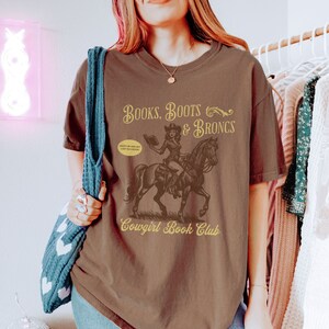 May include: Brown t-shirt with a graphic of a cowgirl riding a horse and holding a book. The text "Books, Boots & Broncs" is above the graphic and "Cowgirl Book Club" is below. The text "Giddy up and get lost in a book" is in a speech bubble.