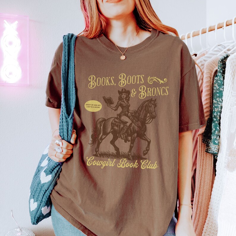 May include: Brown t-shirt with a graphic of a cowgirl riding a horse and holding a book. The text "Books, Boots & Broncs" is above the graphic and "Cowgirl Book Club" is below. The text "Giddy up and get lost in a book" is in a speech bubble.