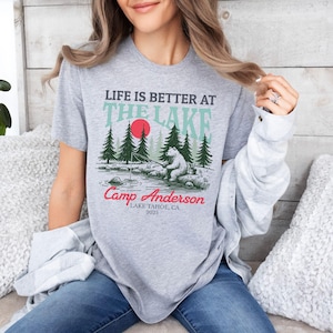 K&ouml;nnte beinhalten: Ein graumeliertes T-Shirt mit einer Grafik eines B&auml;ren, der an einem See angelt, umgeben von Tannen und einer roten Sonne. Der Text lautet: LIFE IS BETTER AT THE LAKE, Camp Anderson, LAKE TAHOE, CA, 2025. Das Design verwendet schwarze, t&uuml;rkise und rote Tinte.