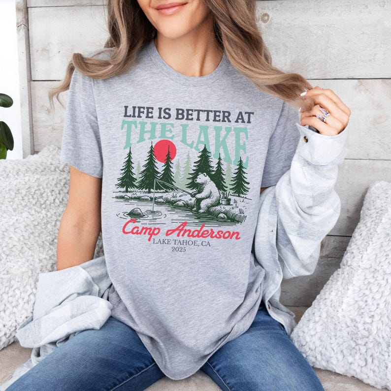K&ouml;nnte beinhalten: Ein graumeliertes T-Shirt mit einer Grafik eines B&auml;ren, der an einem See angelt, umgeben von Tannen und einer roten Sonne. Der Text lautet: LIFE IS BETTER AT THE LAKE, Camp Anderson, LAKE TAHOE, CA, 2025. Das Design verwendet schwarze, t&uuml;rkise und rote Tinte.