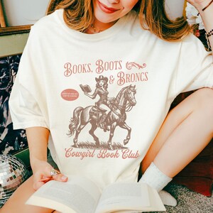 May include: A white t-shirt with a vintage-style graphic design featuring a cowgirl riding a horse and reading a book. The text on the shirt reads "BOOKS, BOOTS & BRONCS" and "Cowgirl Book Club".