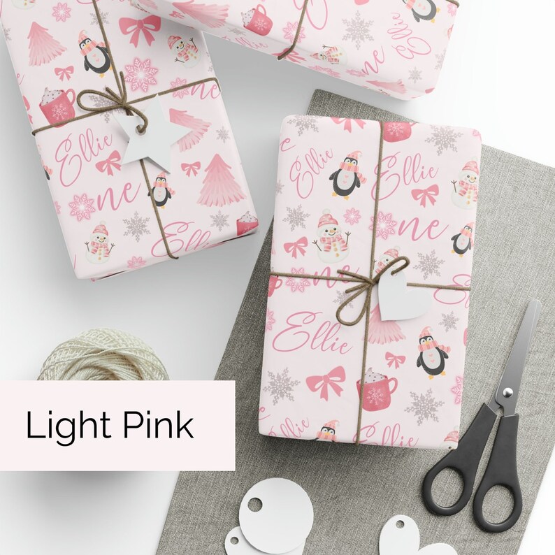 May include: Pink wrapping paper with a repeating pattern of penguins, snowmen, and hot chocolate mugs. The name "Ellie" is repeated throughout the design.