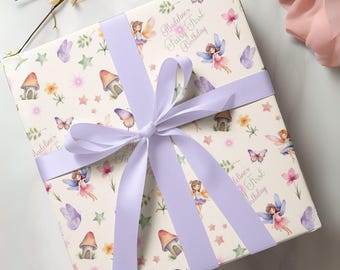 May include: A square gift box wrapped in cream-colored paper featuring a pattern of fairies, mushrooms, butterflies, and stars. The text reads Madeline's Fairy First Birthday. A wide, light purple satin ribbon is tied in a bow around the box.