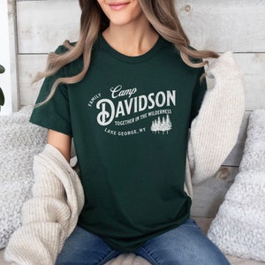 May include: A dark green t-shirt with a white vintage-style graphic that says "Camp Davidson Family Together in the Wilderness Lake George, NY".