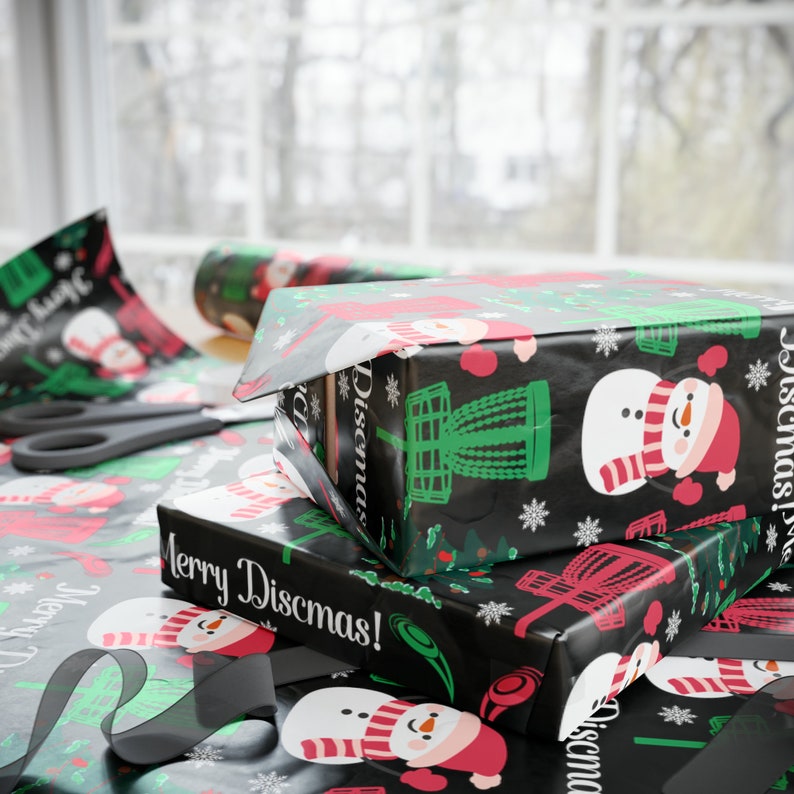 Może przedstawiać: Stos trzech pudełek prezentowych owiniętych w czarno-zielony papier ozdobny z motywem bałwana i kosza do frisbee. Pudełka są oznaczone napisami "Merry Discmas!" i "Discmas!"