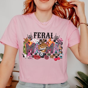 May include: Pale pink t-shirt featuring the words "FERAL MAMA" in a striking design. The graphic includes a tiger, flames, playing cards, and other motifs. The top has short sleeves and a round neckline.