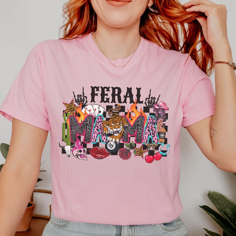 May include: Pale pink t-shirt featuring the words "FERAL MAMA" in a striking design. The graphic includes a tiger, flames, playing cards, and other motifs. The top has short sleeves and a round neckline.