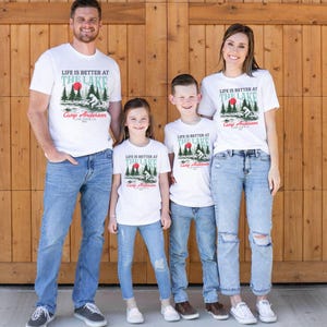 K&ouml;nnte beinhalten: Vier wei&szlig;e T-Shirts mit einer Grafik aus Wald, roter Sonne und B&auml;r sowie dem Text "LIFE IS BETTER AT THE LAKE", "Camp Anderson" und "LAKE TAHOE, CA 2023". Die T-Shirts werden mit blauen Jeans in verschiedenen Ausf&uuml;hrungen kombiniert.