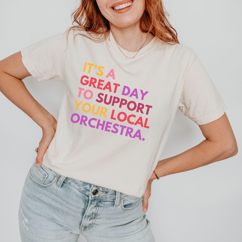 May include: A white t-shirt with the text "It's a great day to support your local orchestra." printed in rainbow colors.