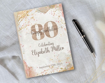 Op de afbeelding: Een rechthoekig gastenboek met een crème en roze aquarelachtergrond, gouden glitteraccenten en de tekst "80 happy birthday Celebrating Elizabeth Miller September 15th, 2024" in bruin handschrift, vergezeld van een zwarte balpen.