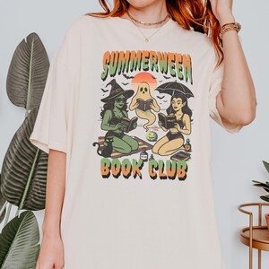 Pode incluir: Camiseta creme com um gr&aacute;fico de tr&ecirc;s figuras lendo livros sob um p&ocirc;r do sol, com as palavras "SUMMERWEEN BOOK CLUB" em verde e laranja. As figuras incluem uma bruxa, um fantasma e uma mulher com um guarda-chuva.