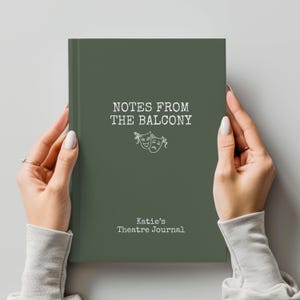 May include: A dark olive green journal with white text that reads NOTES FROM THE BALCONY and Katie's Theatre Journal, featuring a white line drawing of two theatrical masks, one smiling and one frowning, centered on the front cover.