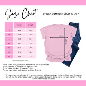 Pode incluir: Uma t-shirt rosa unissexo com uma tabela de tamanhos e o texto "UNISEX COMFORT COLORS 1717". A tabela mostra as medidas do peito e do comprimento em polegadas para os tamanhos S a 4XL. Uma cal&ccedil;a de ganga azul &eacute; parcialmente vis&iacute;vel.