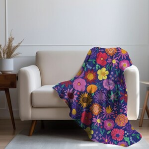 May include: A purple throw blanket with a floral pattern featuring various flowers in shades of red, yellow, pink, and orange. The blanket is draped over a white couch.