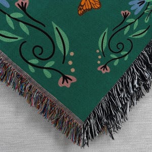 May include: A green woven throw blanket with a black and white fringe border. The blanket features a repeating pattern of butterflies, flowers, and leaves.