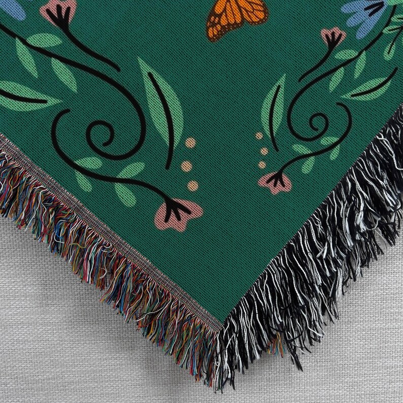 May include: A green woven throw blanket with a black and white fringe border. The blanket features a repeating pattern of butterflies, flowers, and leaves.