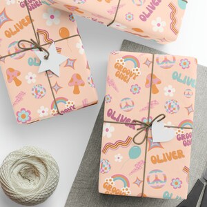 May include: Pink wrapping paper with a groovy 70s theme. The paper features rainbows, peace signs, mushrooms, flowers, and the name "Oliver" repeated throughout. The text "Groovy One!" is also featured on the paper.