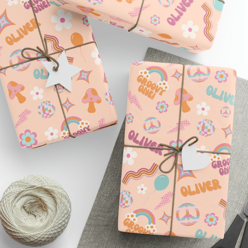 May include: Pink wrapping paper with a groovy 70s theme. The paper features rainbows, peace signs, mushrooms, flowers, and the name "Oliver" repeated throughout. The text "Groovy One!" is also featured on the paper.