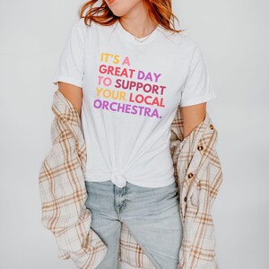 May include: A white t-shirt with the text "It's a great day to support your local orchestra." printed in rainbow colors. The shirt is being worn by a person with a plaid shirt over it.
