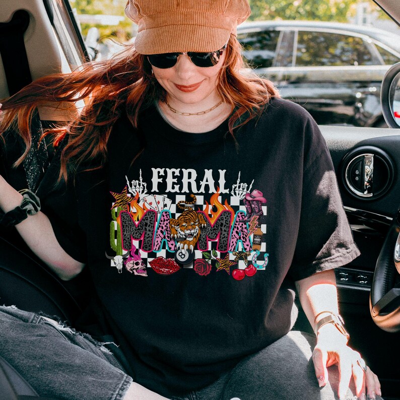 May include: Black t-shirt featuring the text "FERAL MAMA" in a vibrant graphic. The design incorporates a tiger, flames, skulls, and other motifs. The shirt has a relaxed, oversized fit.
