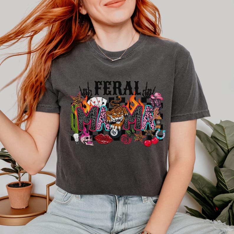 May include: Dark grey t-shirt featuring the words "FERAL MAMA" in a vibrant, multi-coloured design. The design incorporates a tiger, flames, and other illustrations. The t-shirt has a crew neck and short sleeves.