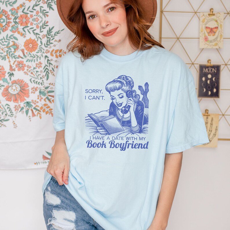 Op de afbeelding: Een lichtblauw T-shirt met een retro-illustratie van een vrouw die een boek leest en aan de telefoon praat. De tekst op het shirt luidt "Sorry, I can't. I have a date with my Book Boyfriend."