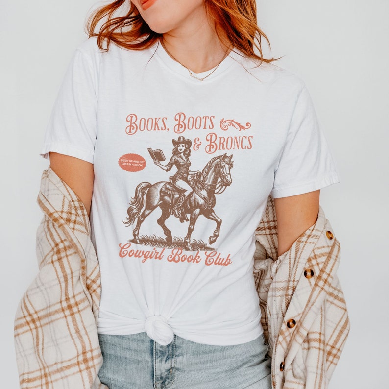 May include: White t-shirt with a graphic of a woman riding a horse and reading a book. The text on the shirt reads "Books, Boots & Broncs Cowgirl Book Club".