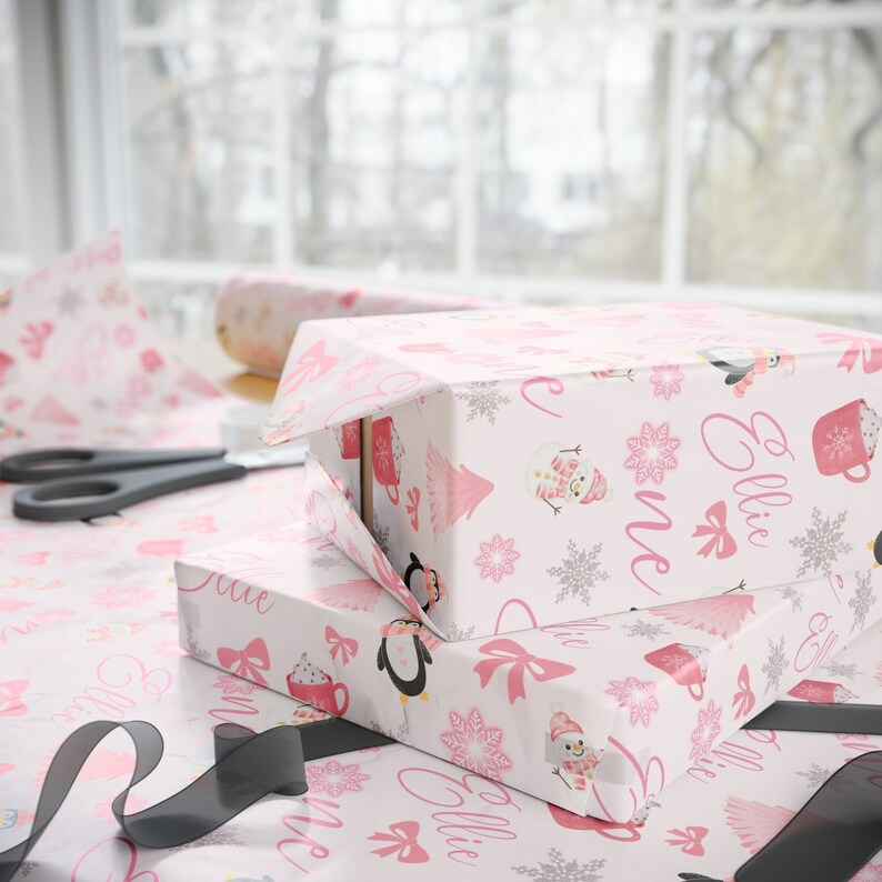May include: Two pink and white gift boxes with a winter theme. The boxes are wrapped in paper with a snowflake pattern and feature penguins, snowmen, and the name "Ellie" written in pink.