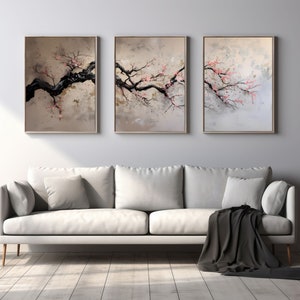 Może przedstawiać: Trzy oprawione abstrakcyjne dzieła sztuki przedstawiające czarne gałęzie drzew z różowymi kwiatami na neutralnym tle. Grafika jest eksponowana nad białą sofą z szarymi poduszkami i czarnym kocem. Widoczna jest również lampa podłogowa i roślina doniczkowa.