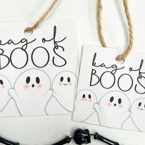 Puede incluir: Dos etiquetas de regalo cuadradas blancas con esquinas redondeadas, cada una con el texto negro "bag of BOOS" y una ilustraci&oacute;n de tres fantasmas blancos con mejillas rosadas. Cada etiqueta tiene un agujero con un cordel marr&oacute;n para colgar.