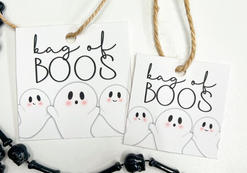 Puede incluir: Dos etiquetas de regalo cuadradas blancas con esquinas redondeadas, cada una con el texto negro "bag of BOOS" y una ilustraci&oacute;n de tres fantasmas blancos con mejillas rosadas. Cada etiqueta tiene un agujero con un cordel marr&oacute;n para colgar.