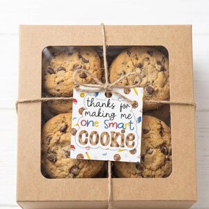 May include: A kraft paper box with a clear window, tied with twine, containing chocolate chip cookies. A tag reads "thanks for making me one smart cookie". The text "Just Print &bull; Cut &bull; Attach" is at the top.