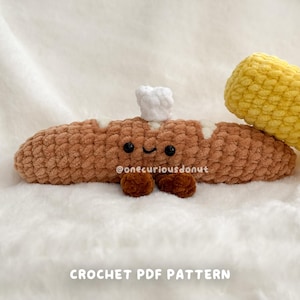 Könnte beinhalten: Ein handgehäkeltes braunes Hotdog mit weißen Akzenten, schwarzen Augen und einem lächelnden Gesicht. Obenauf sitzt eine kleine weiße Kochmütze. Der Text "@onecuriousdonut" und "CROCHET PDF PATTERN" sind ebenfalls sichtbar.