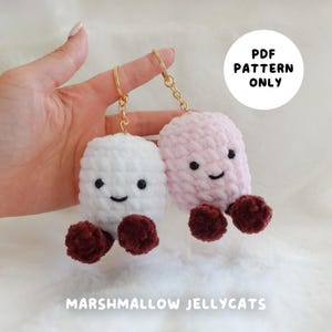 May include: PDF pattern for crocheting two marshmallow-shaped keychains. One keychain is white with a pink nose and the other is pink with a white nose. Both keychains have brown feet and a smiling face.