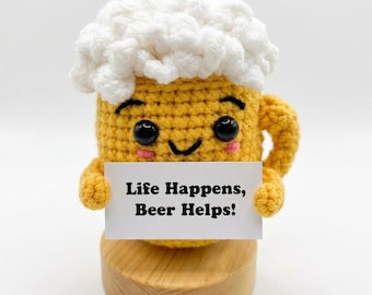 May include: A yellow crocheted beer mug plush with white foam on top, black safety eyes, a stitched smile, and pink cheeks. It holds a white rectangular sign with the black text "Life Happens, Beer Helps!" and sits on a round wooden base.