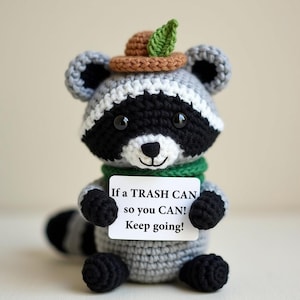 Könnte beinhalten: Gehäkelte Waschbärpuppe mit schwarz-weißem Gesicht, grauem Körper und einem kleinen braunen Hut mit einem grünen Blatt. Der Waschbär hält ein Schild mit der Aufschrift: "If a TRASH CAN so you CAN! Keep going!"