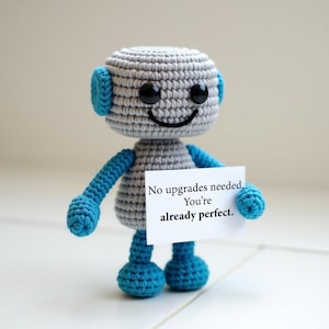 May include: Crocheted robot toy in gray and blue, holding a white sign. The robot has a smiling face with black eyes and a black smile. The sign reads, "No upgrades needed. You're already perfect."