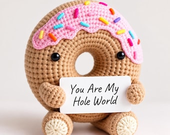 May include: A tan crocheted donut plush with a pink icing top and colorful sprinkles. It features two small black eyes, two arms, and two legs. The donut holds a white rectangular sign with the black text "You Are My Hole World" printed on it.