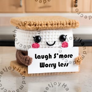 Puede incluir: Un juguete de peluche de s'more tejido a crochet que sostiene un cartel que dice "Laugh S'more Worry Less". El s'more está hecho de hilo marrón, blanco y beige.
