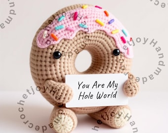 May include: A crocheted donut toy holding a card that says "You Are My Hole World". The donut is brown with pink frosting and colorful sprinkles.