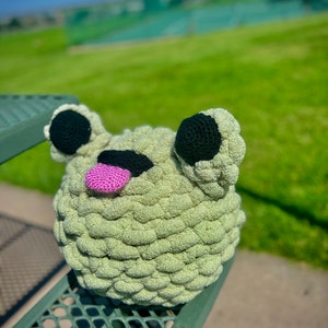 May include: A green crocheted frog toy with black eyes and a pink tongue. The frog is sitting on a green metal bench.