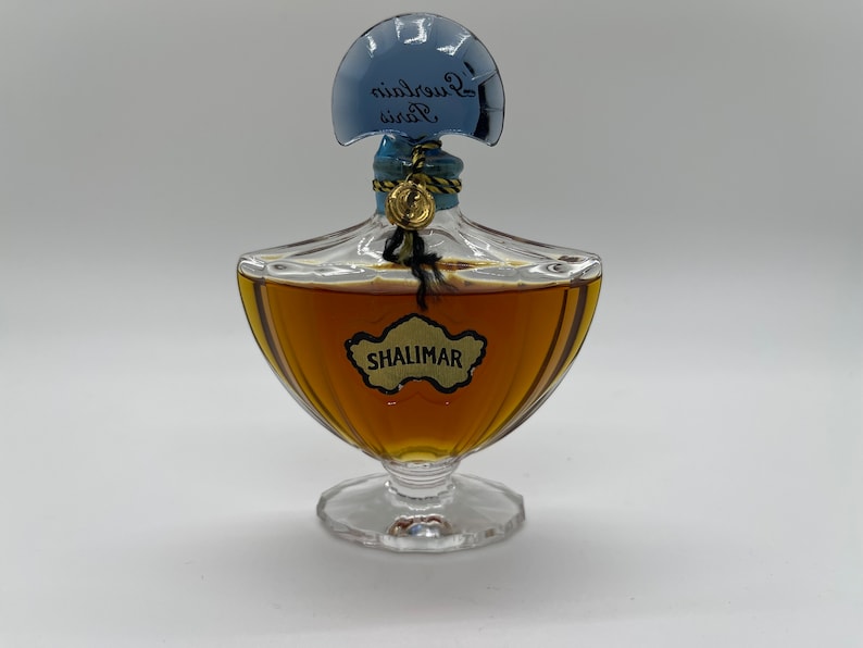 May include: A vintage Shalimar perfume bottle. The clear glass bottle has a unique shape with a blue cap and gold accents. The bottle is filled with a golden-brown liquid, and the label reads "SHALIMAR".
