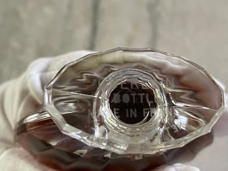 May include: Close-up of a clear, faceted perfume bottle with a brown liquid inside. The bottle has a geometric design and the text "BOTTLE MADE IN FRANCE" is visible. The bottle is likely vintage.