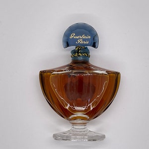 May include: A vintage perfume bottle with a unique shape. The bottle is filled with a brown liquid and has a blue cap with the words "Guerlain Paris" in gold lettering. The bottle sits on a clear, decorative base.