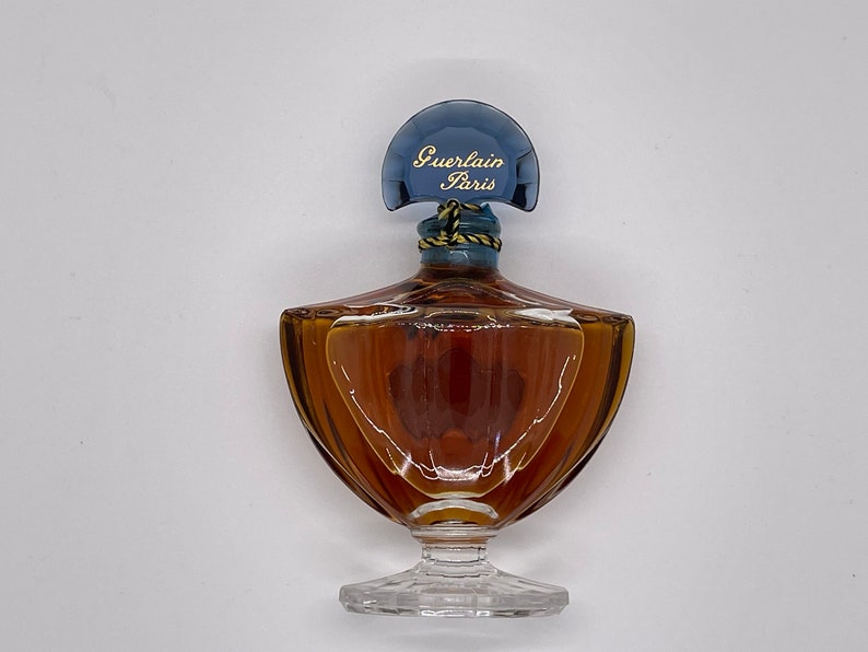 May include: A vintage perfume bottle with a unique shape. The bottle is filled with a brown liquid and has a blue cap with the words "Guerlain Paris" in gold lettering. The bottle sits on a clear, decorative base.