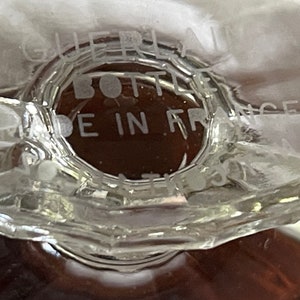May include: Close-up of a clear glass bottle base with the text "GUERLAIN BOTTLE MADE IN FRANCE". The bottle has a geometric design and a brown liquid inside.